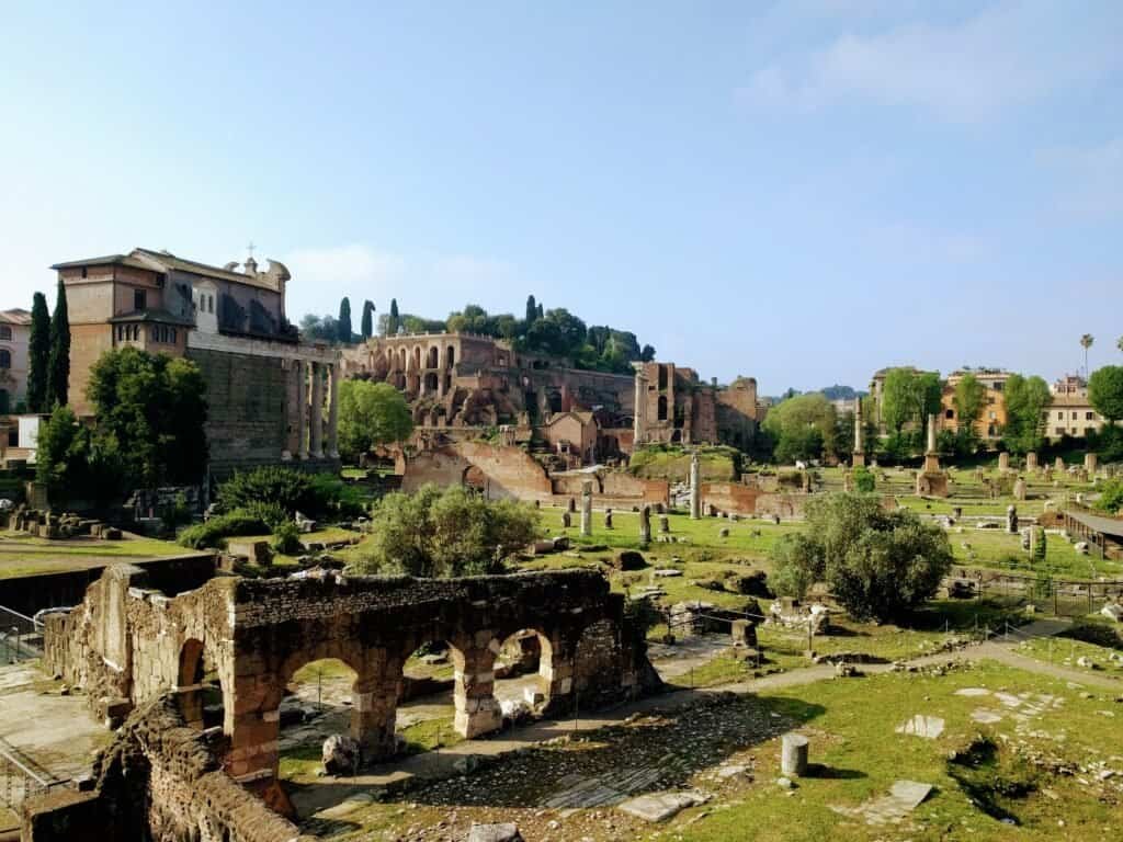 Ruins of ancient Roman structures with historic buildings and greenery in the background, showcasing Rome's archaeological heritage.