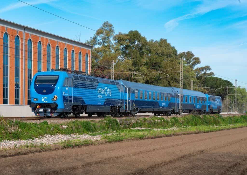 A sleek blue electric train traveling along the railway tracks near a cityscape with a brick building and green trees in the background. Getting the train is one of our Italy travel tips - it's the perfect way to travel.