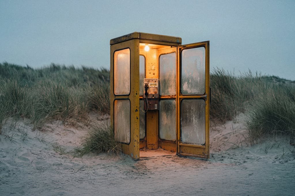 An isolated vintage phone booth on a sandy beach with grass dunes, illuminated from within during twilight, creating a nostalgic and tranquil seaside scene.