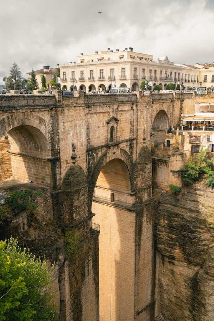 Ancient stone bridge with arches spanning a deep gorge, featuring a historic building on top, under a cloudy sky.